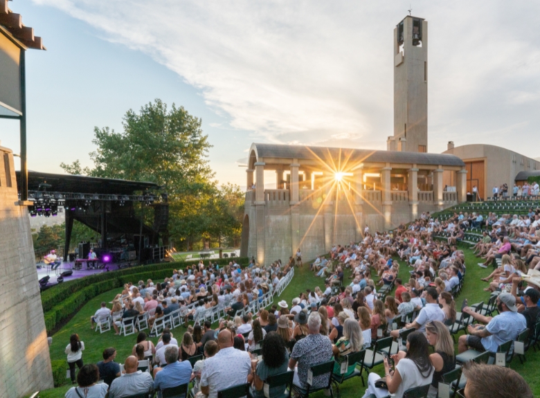 Guests gather in the outdoor amphitheatre at Mission Hill Family Estate Winery in West Kelowna during the Summer Concert Series, with the iconic bell tower and estate grounds illuminated by the evening sun.