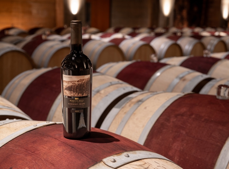 A bottle of Mission Hill Terroir Cabernet Franc rests atop oak barrels in the estate barrel cellar, highlighting the craftsmanship behind premium Okanagan wines.